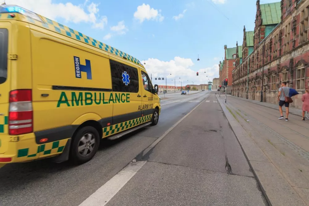 En gul ambulance med grønne ternede markeringer kører ned ad en stort set tom bygade i nærheden af historiske bygninger i europæisk stil; to fodgængere går på fortovet.