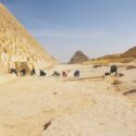 En gruppe mennesker arbejder ved foden af store, gamle pyramider i et sandet, solrigt område med stenblokke og arkæologiske redskaber spredt ud over jorden.