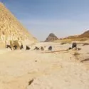 En gruppe mennesker arbejder ved foden af store, gamle pyramider i et sandet, solrigt område med stenblokke og arkæologiske redskaber spredt ud over jorden.