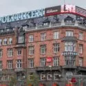 En historisk murstensbygning med store blå POLITIKEN-skilte og elektroniske billboards på taget, beliggende på en travl gade med lidt grønt og en rød metroindgang foran.