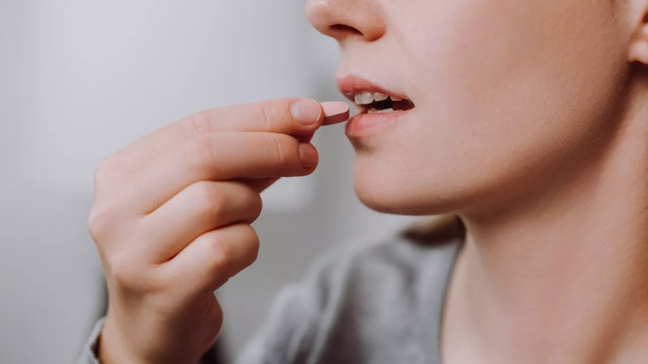 Nærbillede af en person, der holder en lyserød D-vitamintablet tæt på munden og er ved at tage pillen. Personen er iført en grå skjorte, og kun den nederste halvdel af ansigtet og hånden er synlig.