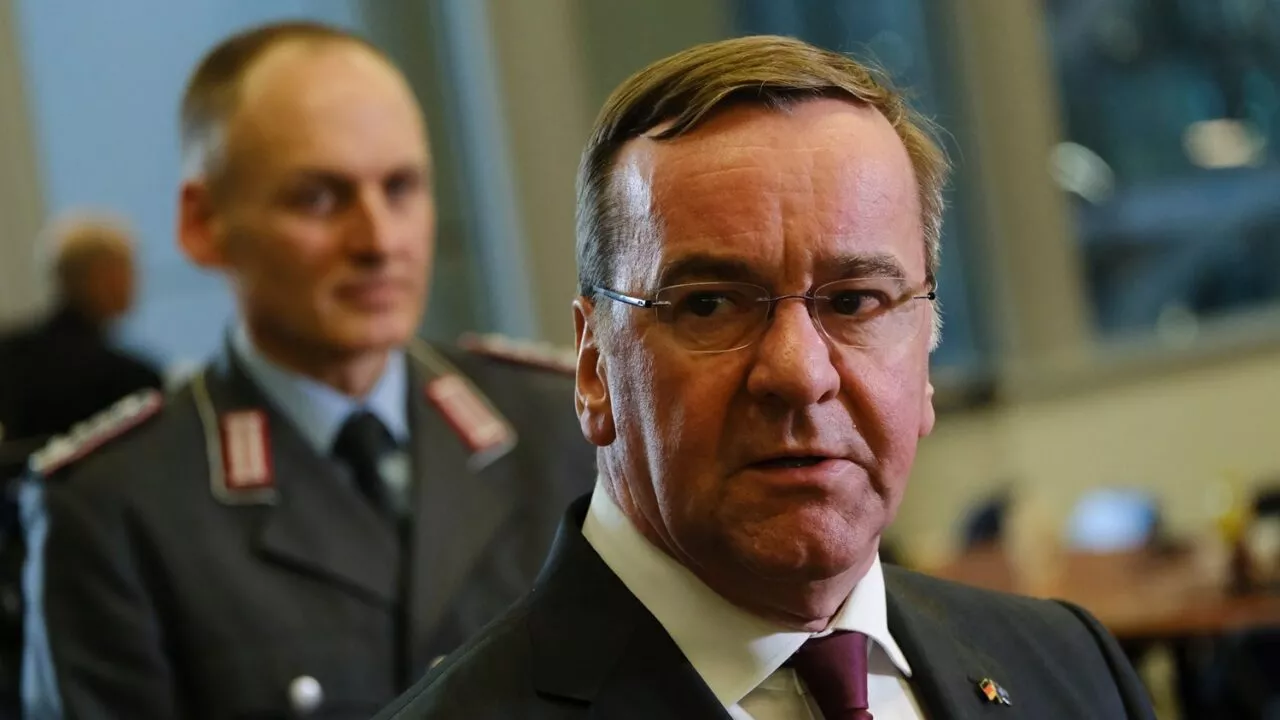 En mand i jakkesæt og briller står i fokus med et alvorligt udtryk, mens en anden mand i militæruniform står sløret i baggrunden. De ser ud til at være indendørs, muligvis til en formel begivenhed eller et møde.