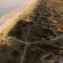 Luftfoto af en sandkyst med bølgende klitter ud til et roligt hav. Spredte huse og snoede veje er synlige mellem klitterne, og det varme sollys kaster lange skygger over landskabet.