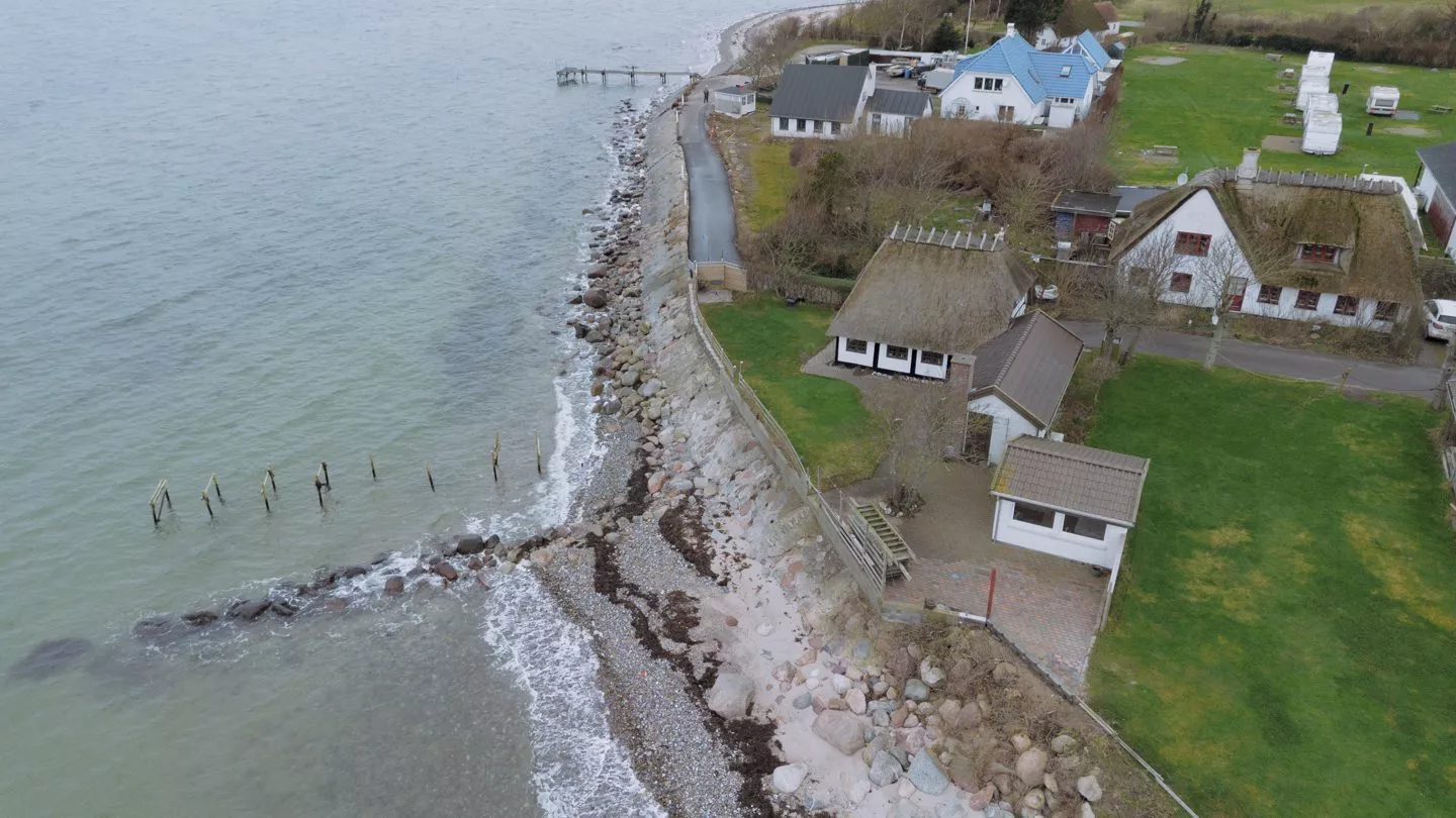 Luftfoto af en kystby med huse nær kysten, en stenet strand, grønne græsplæner, en smal vej og en træmole, der strækker sig ud over vandet.