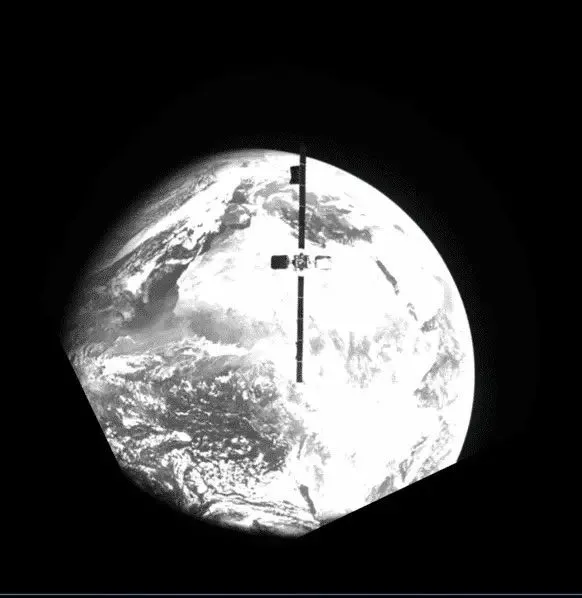 Et sort-hvidt billede af Jorden fra rummet med en satellit i silhuet foran planeten, som delvist dækker udsigten, og en sort kant i nederste venstre hjørne.