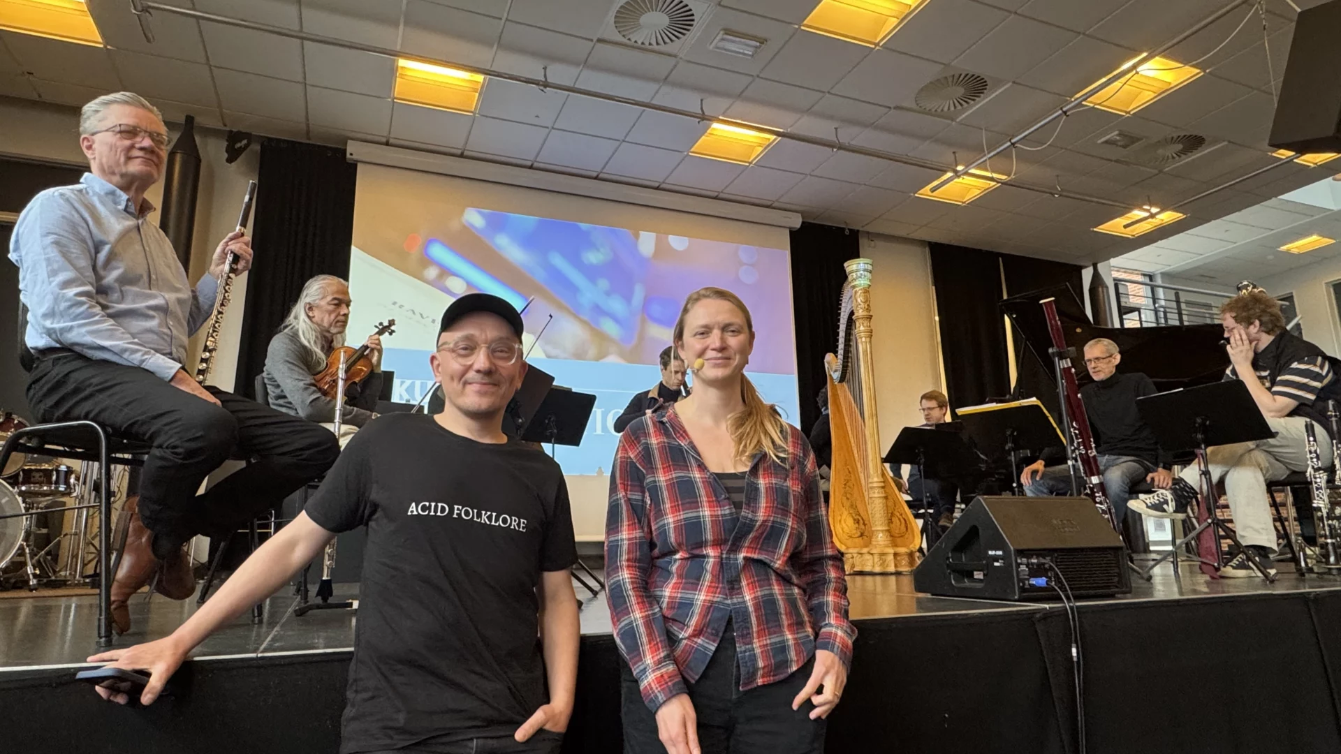 En gruppe musikere med instrumenter sidder og står på en scene indendørs. To personer, den ene i sort skjorte og kasket, den anden i ternet skjorte, poserer og smiler foran. En harpe og et projiceret billede ses i baggrunden.