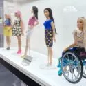 Fem Barbie-dukker er udstillet i en glasmontre. Fire står og er klædt i farverige outfits, mens en dukke sidder i en blå kørestol iført en stribet skjorte og smiler. Baggrunden er hvid og moderne.