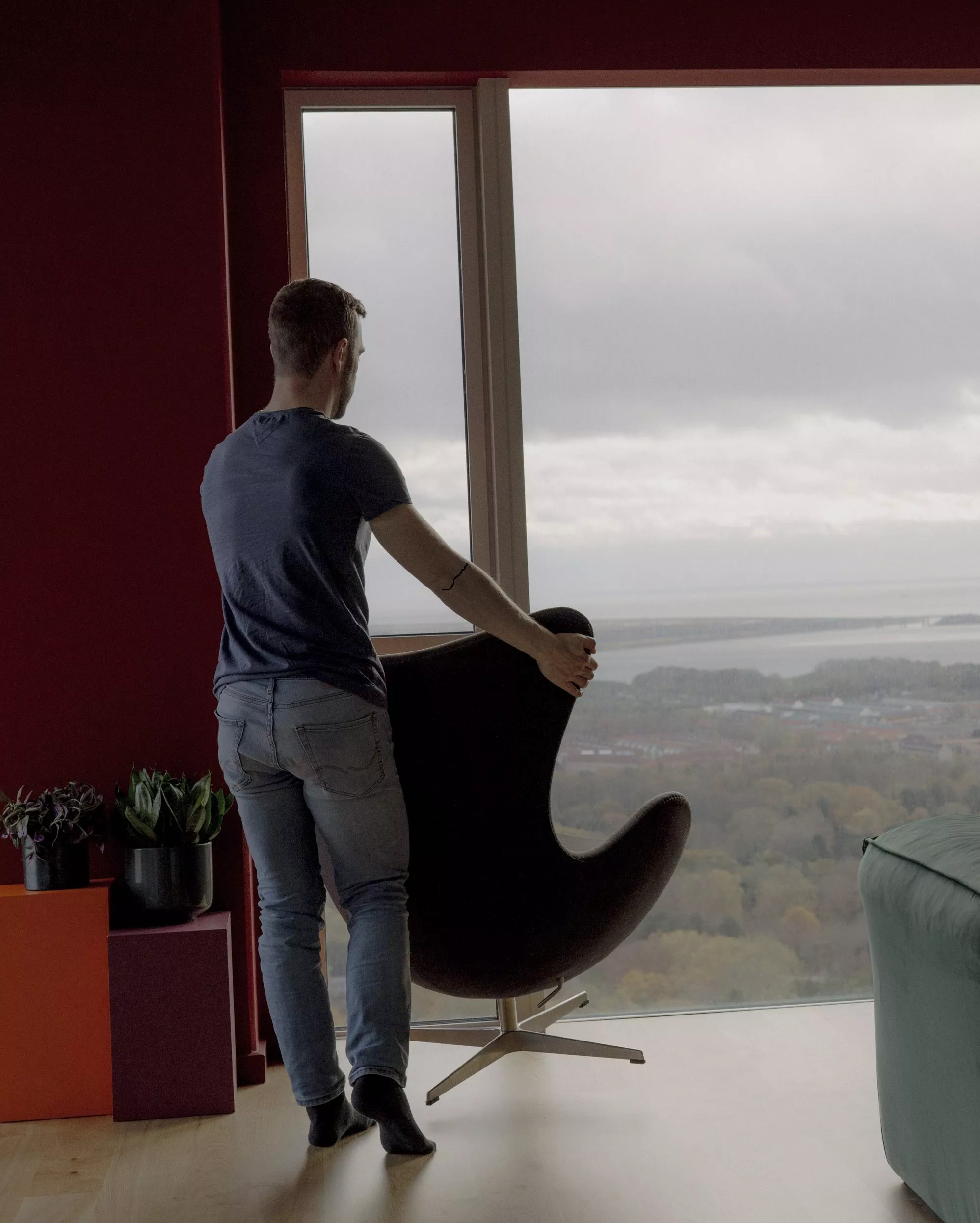 En mand står barfodet i et moderne rum foran et stort vindue med udsigt til træer og vand. Han holder en sort stol, og potteplanter står på farverige stativer i nærheden. Himlen udenfor er overskyet.