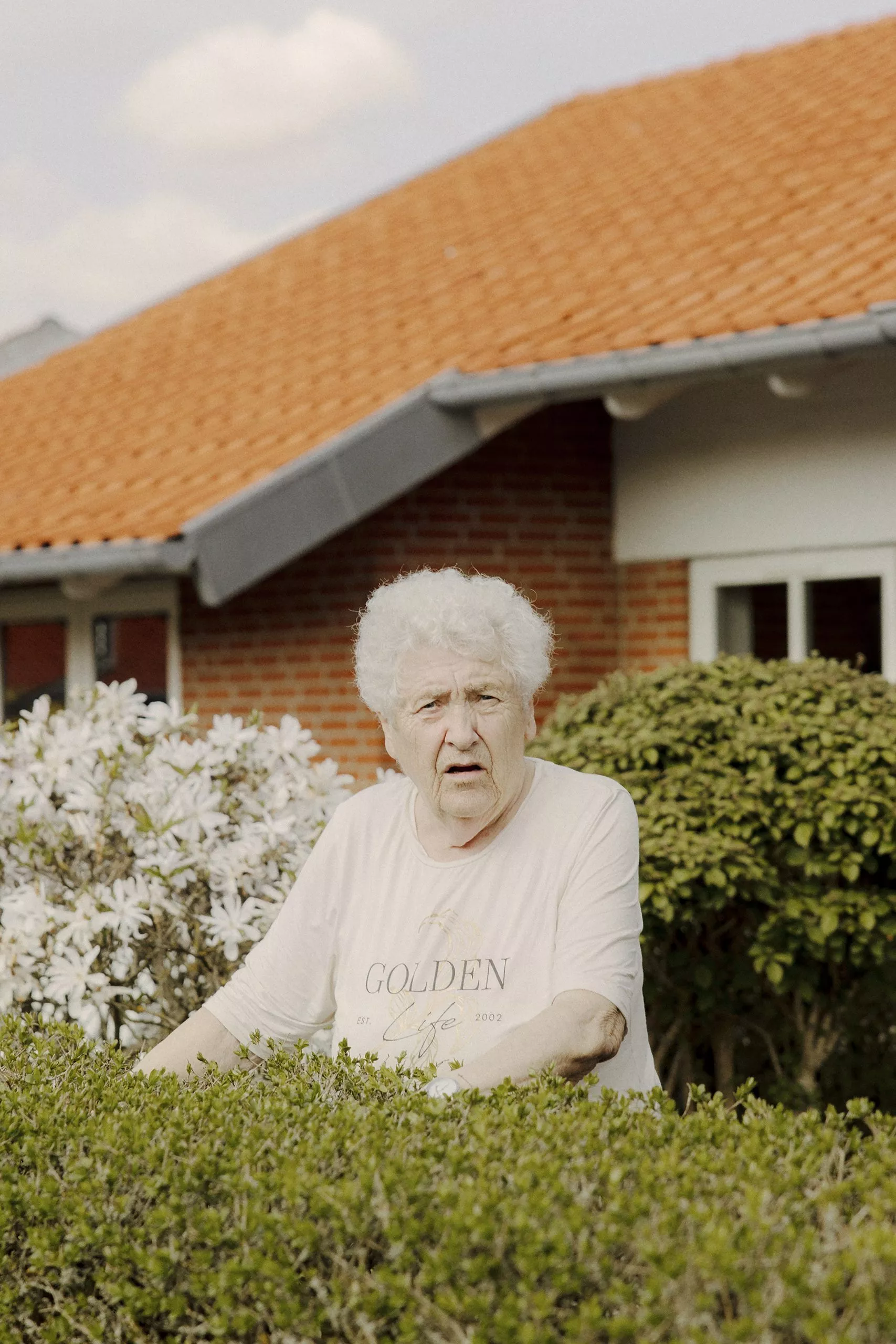 En ældre kvinde med kort hvidt hår står bag grønne buske foran et hus med rødt tegltag, iført en lys GOLDEN T-shirt, med blomstrende planter og blå himmel i baggrunden.