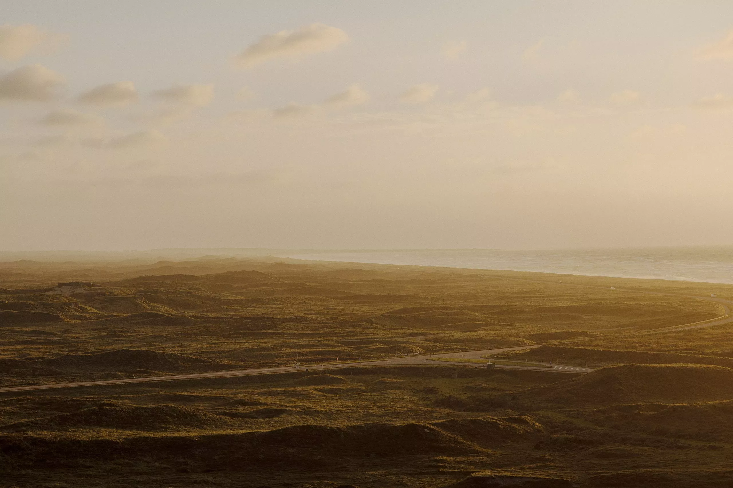 Et kystlandskab ved solnedgang med bølgende græsklædte klitter, en vej, der skærer sig gennem terrænet, og havet, der er synligt til højre under en delvist overskyet himmel. Scenen er badet i et varmt, gyldent lys.
