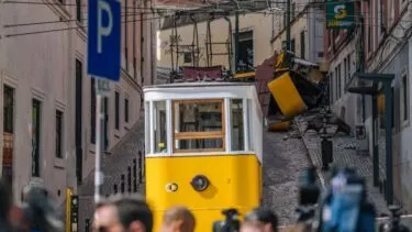 En gul sporvogn holder på stejle brostensskinner i byen nær Elevador da Glória; bag den ser en anden sporvogn ud til at være afsporet og beskadiget. Mennesker og kameraer er synlige i forgrunden, og der er bygninger på begge sider af gaden.