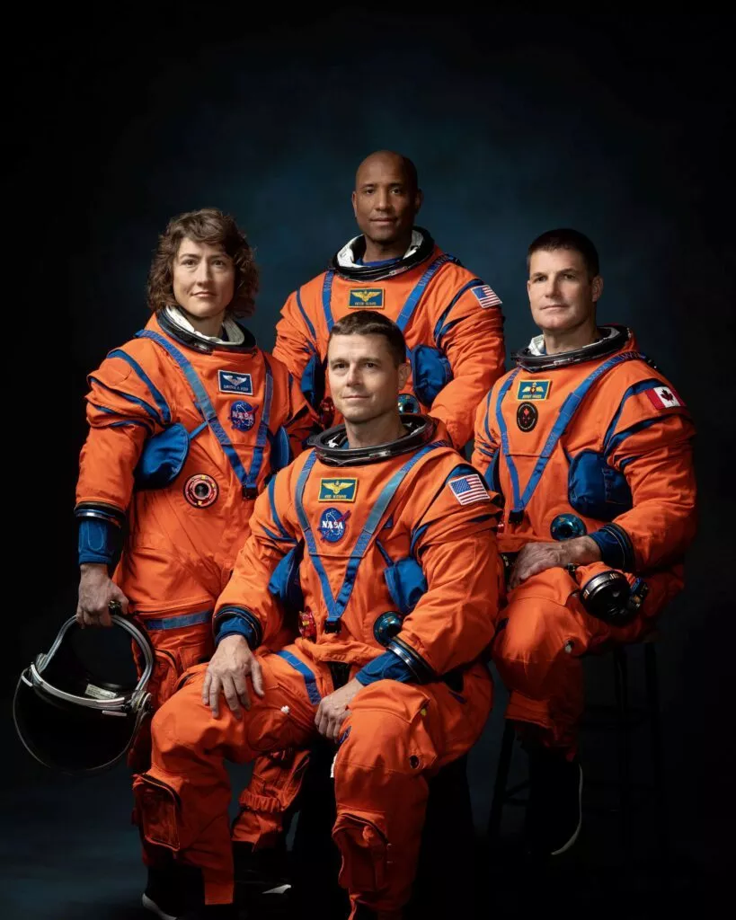 Fire astronauter i lysende orange NASA-rumdragter poserer til et formelt gruppeportræt på en mørk baggrund. Tre står, en sidder, og to holder hjelme. Den ene astronauts dragt har et mærke med det canadiske flag.