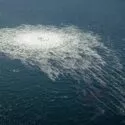 Luftfoto af havet med et stort cirkulært område med boblende vand, sandsynligvis forårsaget af gas eller luft, der slipper ud under overfladen og skaber en skummende forstyrrelse midt i de ellers rolige bølger.
