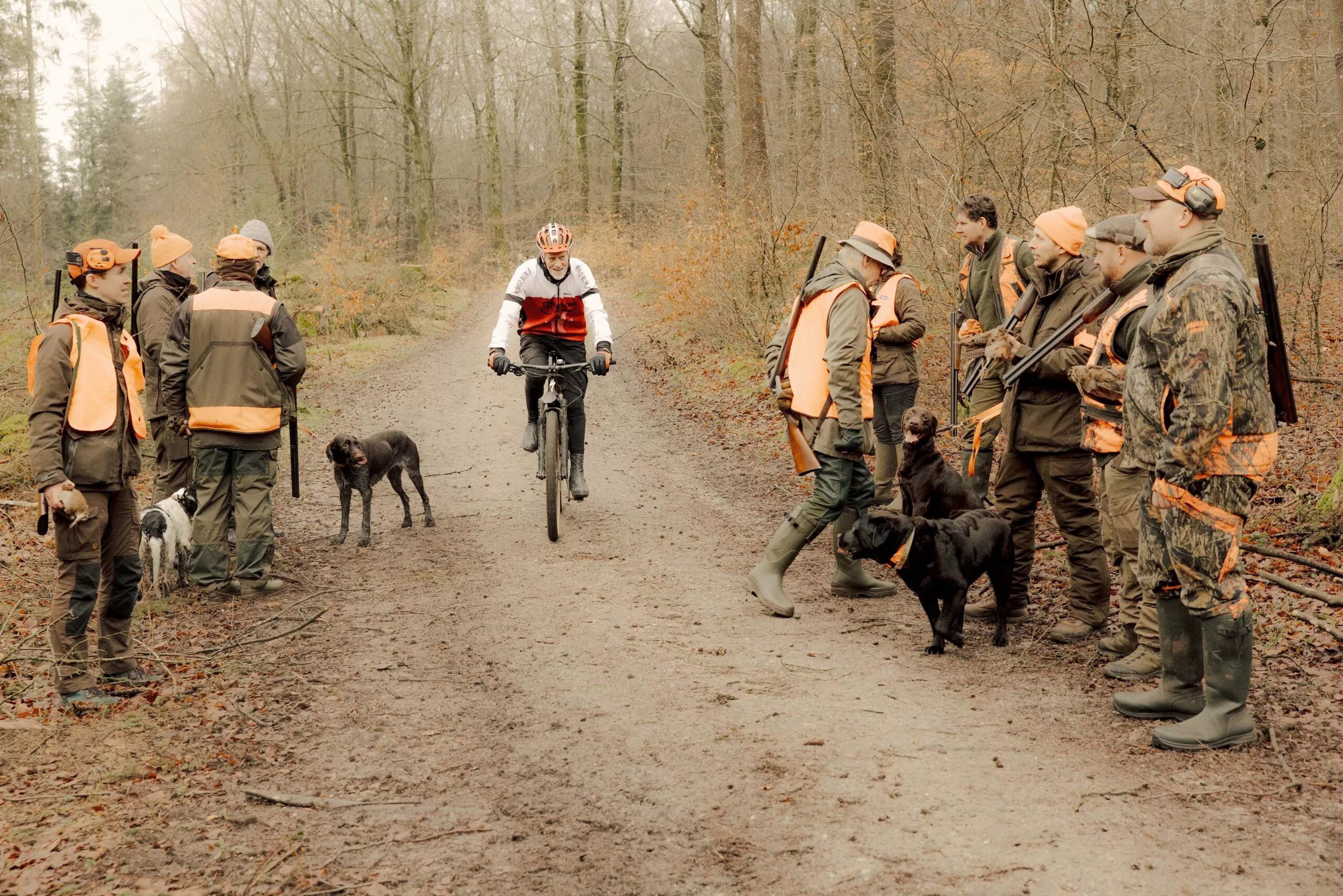 En cyklist kører ned ad en skovsti, mens en gruppe jægere med hunde og rifler, klædt i orange og camouflageudstyr, står ved siden af stien. Scenen udspiller sig på en overskyet efterårs- eller vinterdag.