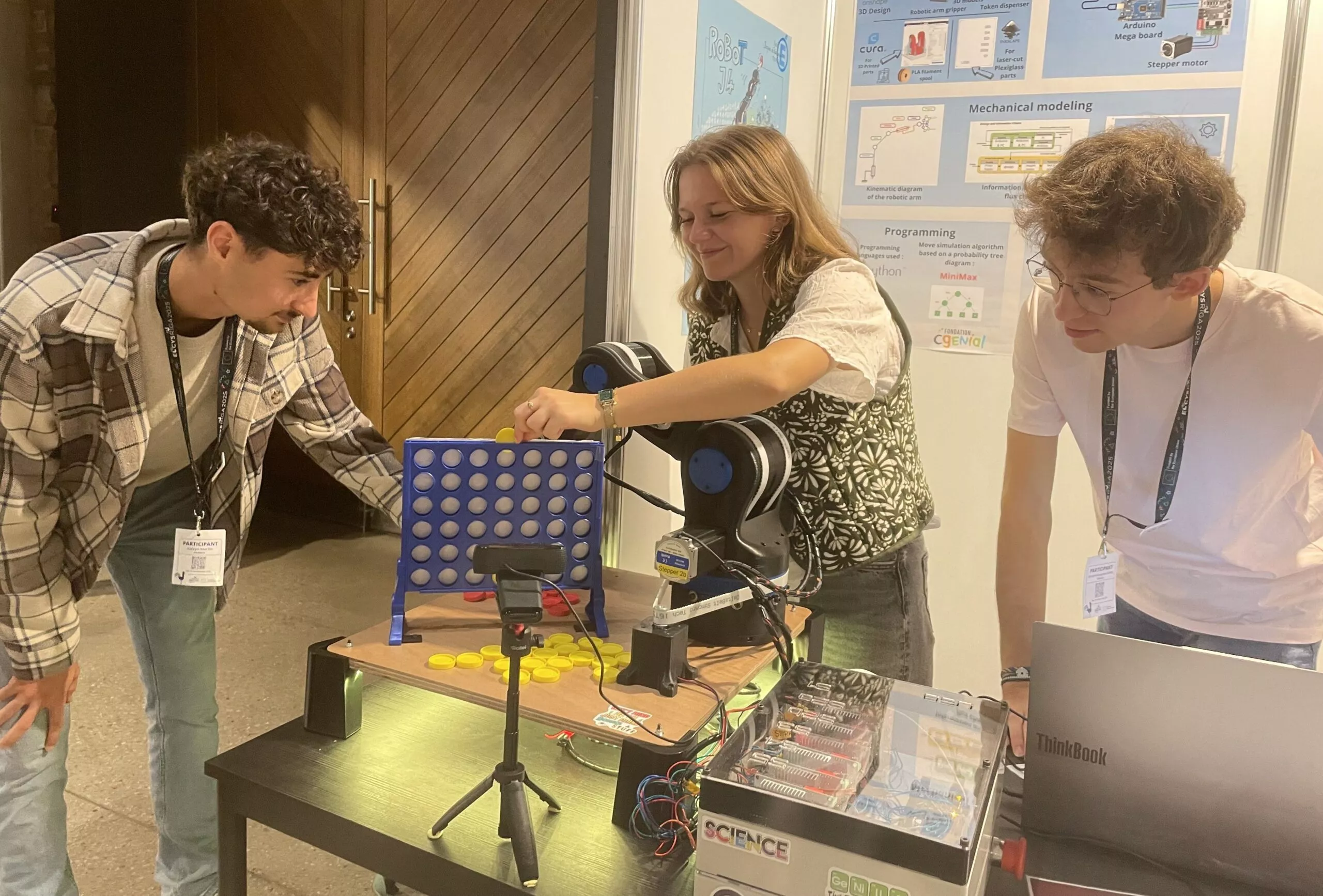 Tre unge mennesker demonstrerer en robotarm, der spiller Connect 4 på en videnskabsmesse. En person justerer robotten, mens en anden smiler og observerer. På bordet ses plakater, en bærbar computer og elektronik.