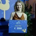 En ung kvinde med krøllet blondt hår smiler, mens hun holder et skilt med teksten Third Prize €3500 ved Riga ELCYS 2025-arrangementet. Hun står foran et blåt bagtæppe med en hane og dekorative planter.
