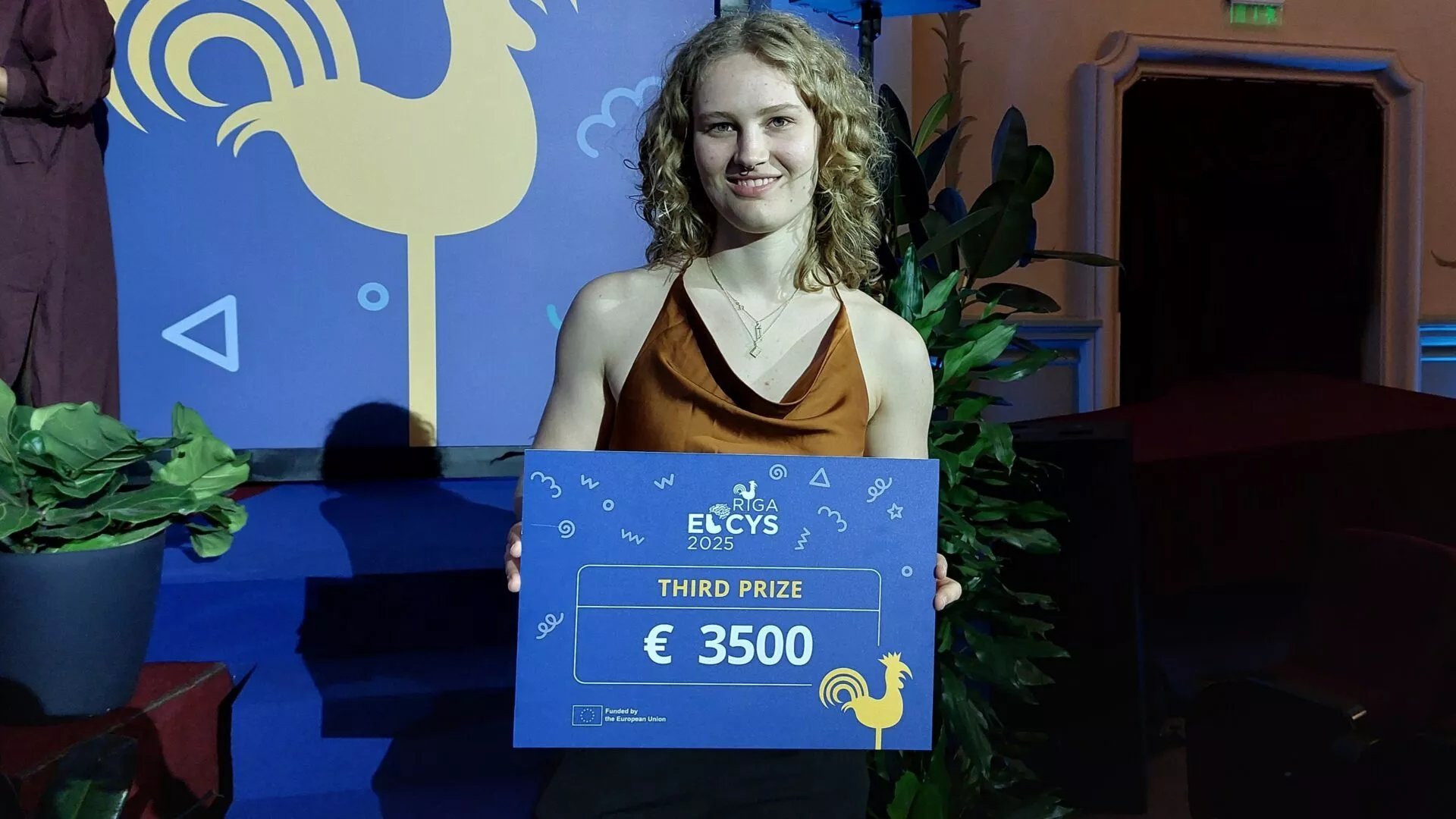 En ung kvinde med krøllet blondt hår smiler, mens hun holder et skilt med teksten Third Prize €3500 ved Riga ELCYS 2025-arrangementet. Hun står foran et blåt bagtæppe med en hane og dekorative planter.