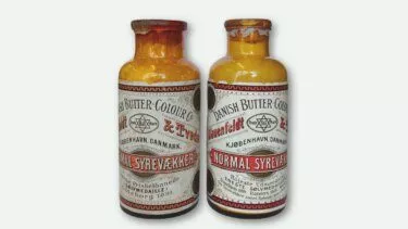 To vintage gule glasflasker mærket Danish Butter Colour med sort, rød og hvid tekst, der viser et stjernesymbol og dansk skrift. Begge flasker har slidte etiketter og brune kapsler.