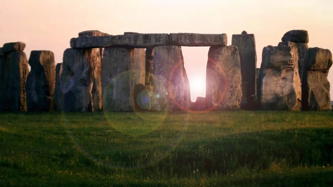 Stonehenges gamle stensøjler står på en græsslette ved solnedgang, og sollyset skinner gennem den centrale bue, så man kan se en overstråling i billedet.