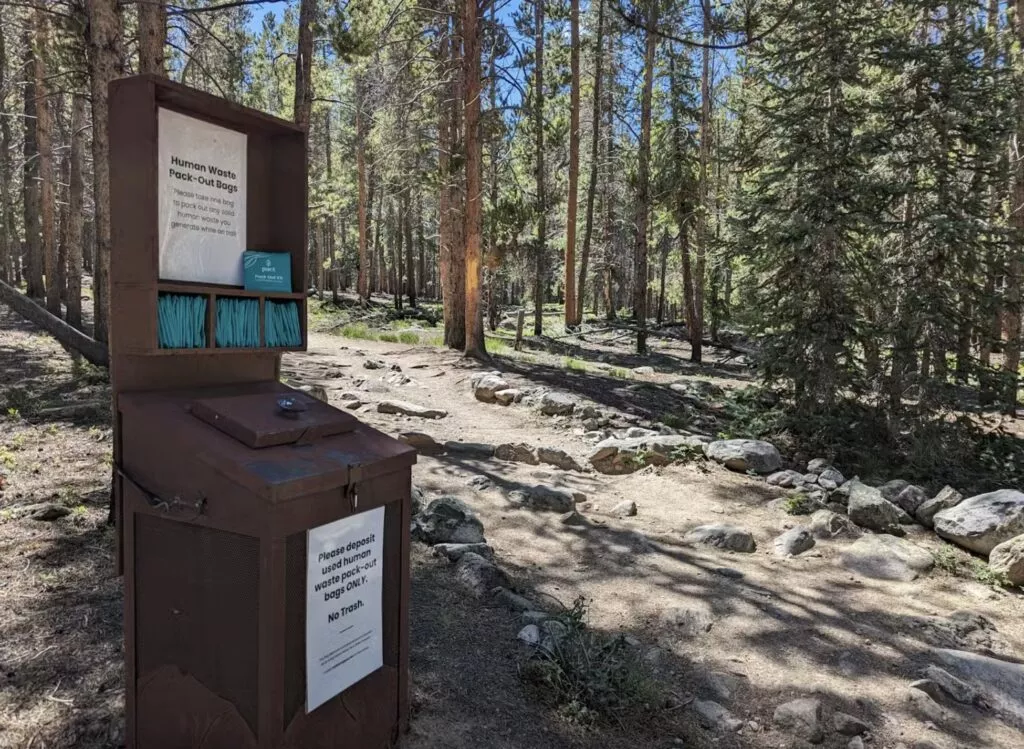 En bjørnesikker container med gratis wag bags til vandrere ved Mount Elbert-ruten nær Leadville og informationsskilte står ved siden af en grussti i en solbeskinnet fyrreskov i Colorado og hjælper poserne bruges til at håndtere menneskelig afføring.