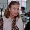 En midaldrende kvinde med skulderlangt lysebrunt hår, briller på hovedet, en beige vest og en top med leopardprint står på et kontor med slørede mennesker og skriveborde i baggrunden.