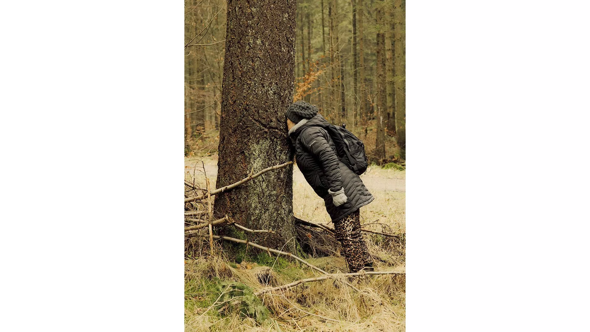 En person i grå frakke, strikhue, handsker og rygsæk står i en skov og læner hovedet mod et stort træ. Træet er omgivet af tørt græs og bladløse grene.