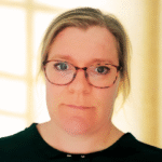 En kvinde med lys hud, iført briller og en sort top, ser direkte ind i kameraet. Hun har blondt hår, der er trukket tilbage, og står foran en svagt oplyst, sløret baggrund.