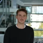 En ung mand med kort brunt hår, iført en sort sweater over en hvid skjorte, står indendørs i en moderne bygning med store vinduer og glasvægge i baggrunden.