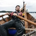 En person i vandtæt overall ror en træbåd på et roligt vand, omgivet af fjerne træer og en overskyet himmel.