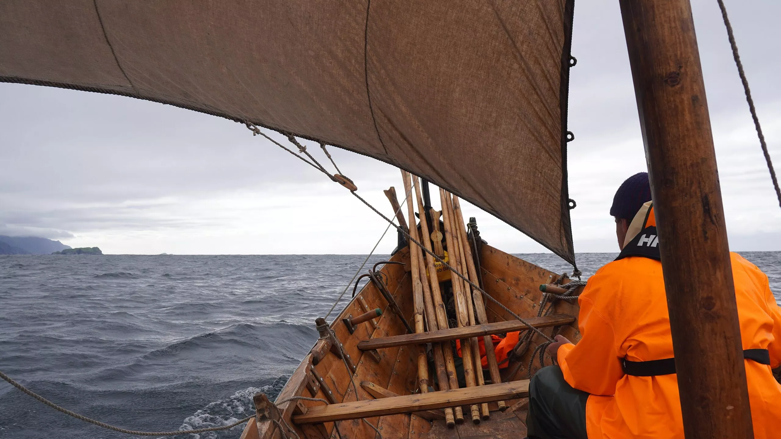 En person i orange jakke sidder i en træsejlbåd på et uroligt hav med træpæle på dækket og et stort, trekantet sejl over hovedet; man kan se en overskyet himmel og et fjernt land.