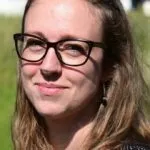En kvinde med langt lysebrunt hår og briller står udendørs og smiler let. Hun er iført en sort og hvid mønstret top, og baggrunden er sløret grønt græs og en hvid bygning.