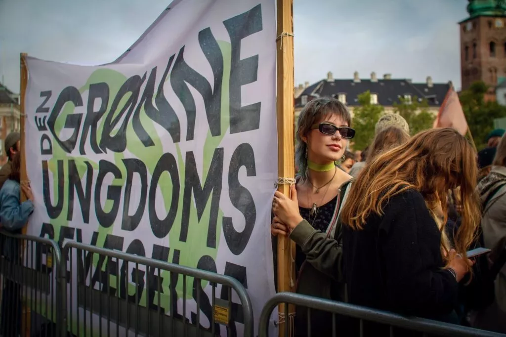 En ung kvinde med solbriller står sammen med andre bag en metalbarriere og holder et stort banner med teksten Den Grønne Ungdomsbevægelse ved en udendørs begivenhed eller protest. Bygninger er synlige i baggrunden.
