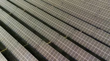 Luftfoto af flere rækker af solpaneler arrangeret i parallelle linjer, der indfanger sollys i en stor solcellepark. Panelerne er placeret på en mørk grund og danner et geometrisk mønster.