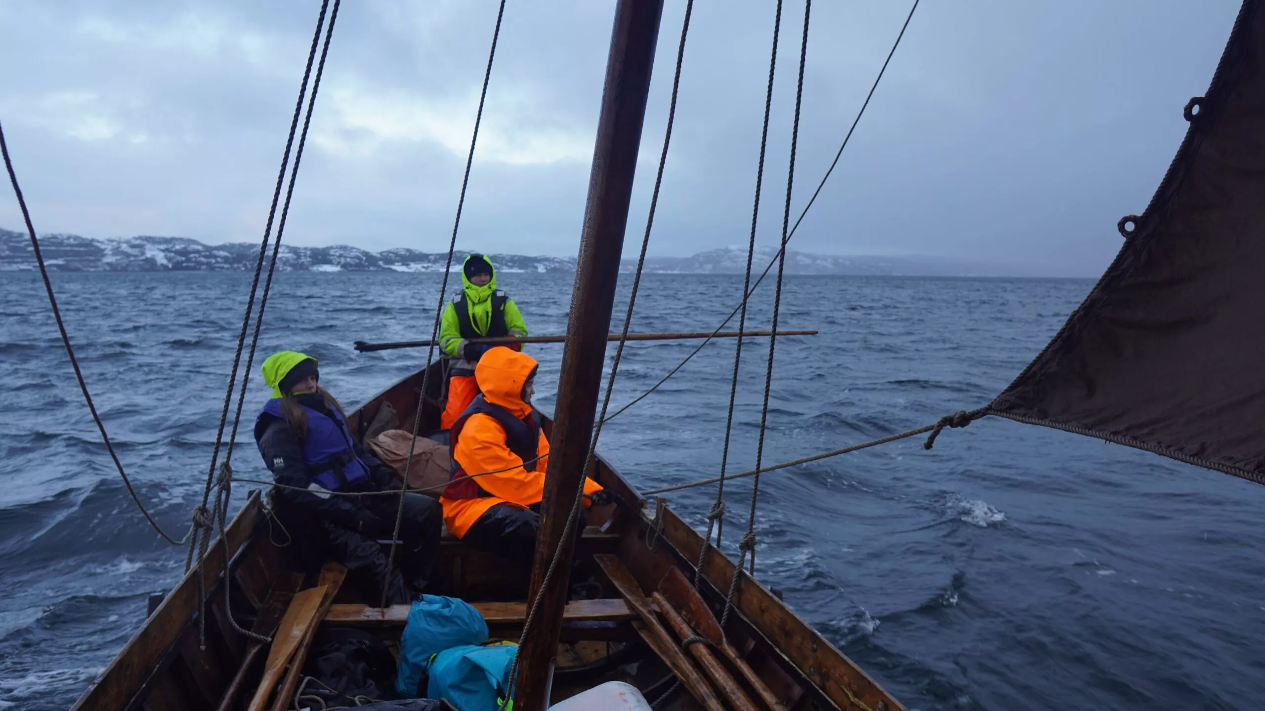 Tre personer iført lyse, vandtætte jakker sidder i en træbåd, der sejler gennem uroligt, koldt vand. Sneklædt, stenet land udgør den fjerne kystlinje under en overskyet himmel.