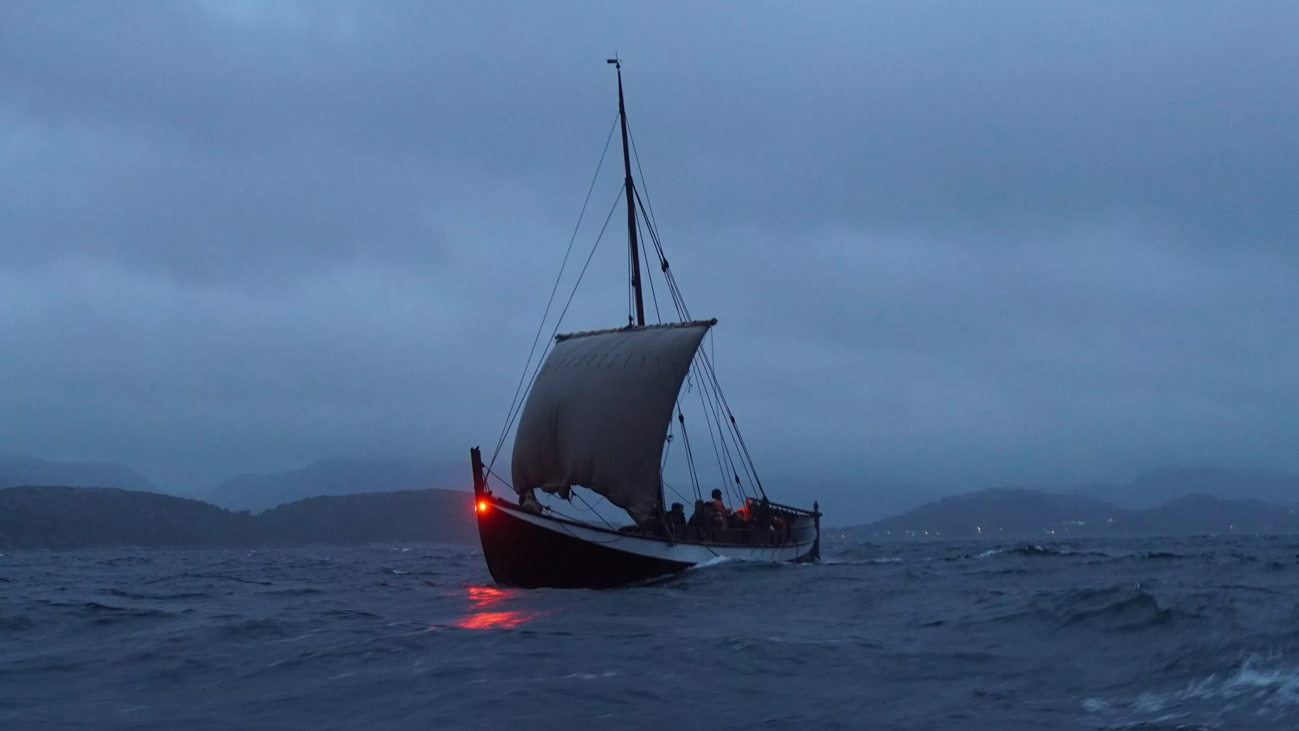 En træsejlbåd med et enkelt sejl og mennesker om bord sejler over uroligt vand i skumringen med bjerge og en overskyet himmel i baggrunden. Et rødt lys lyser bag på båden.