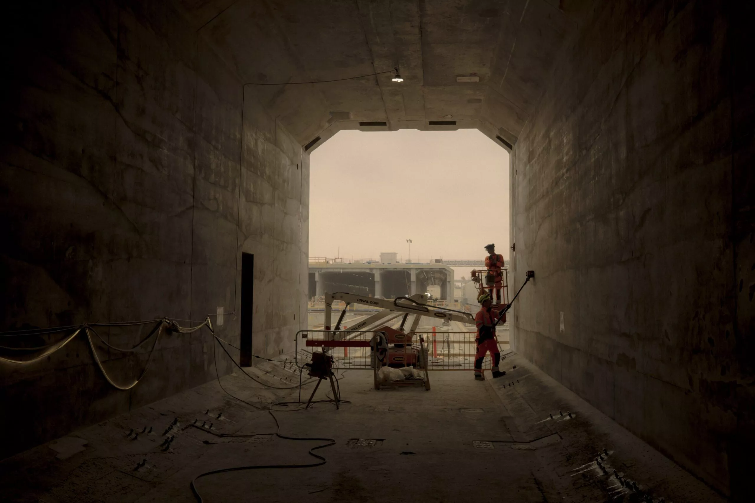 To bygningsarbejdere i orange udstyr arbejder inde i en stor, dunkel betontunnel. Sollyset strømmer ind fra tunnelens ende, hvor man kan se entreprenørmaskiner og en diset himmel udenfor.
