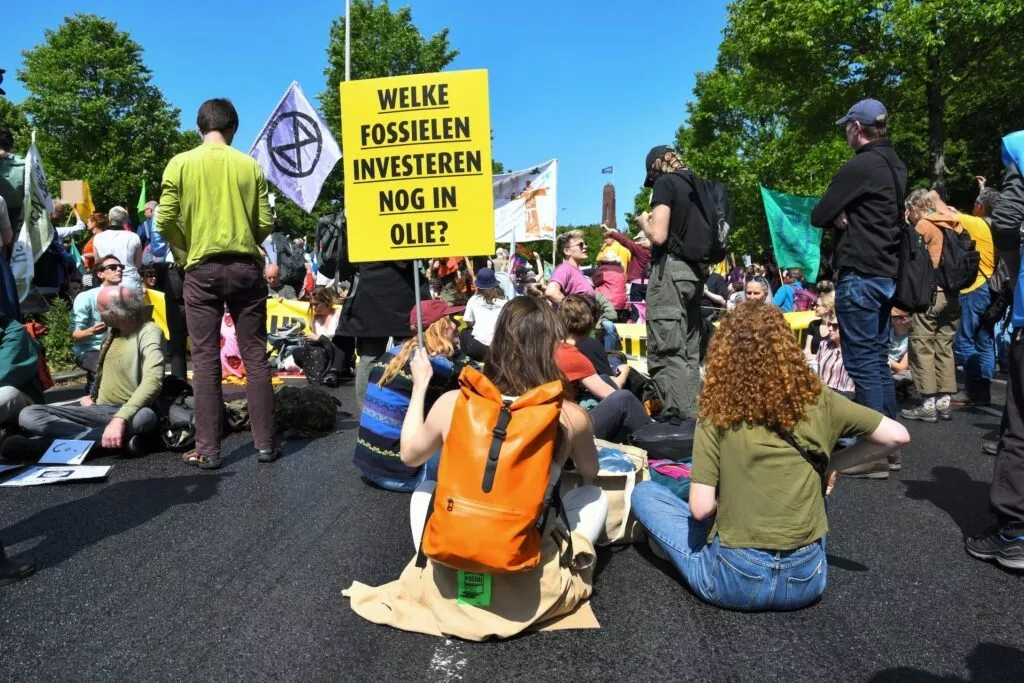En gruppe demonstranter sidder på en gade og holder skilte. På et fremtrædende gult skilt på hollandsk står der: Welke fossielen investeren nog in olie? I baggrunden ses træer og blå himmel.