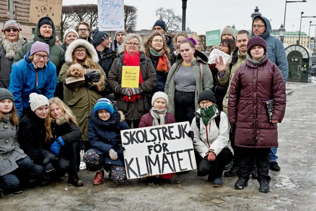 En gruppe mennesker i forskellige aldre står og knæler udendørs i vintertøj med skilte, der støtter klimaindsatsen, herunder et på svensk med teksten Skolstrejk för klimatet (Skolestrejke for klimaet).