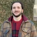 En mand med skæg og langt mørkt hår smiler udendørs iført en brunternet jakke over en rød sweater. Han står foran en mur dækket af efeu med et vindue delvist synligt i baggrunden.