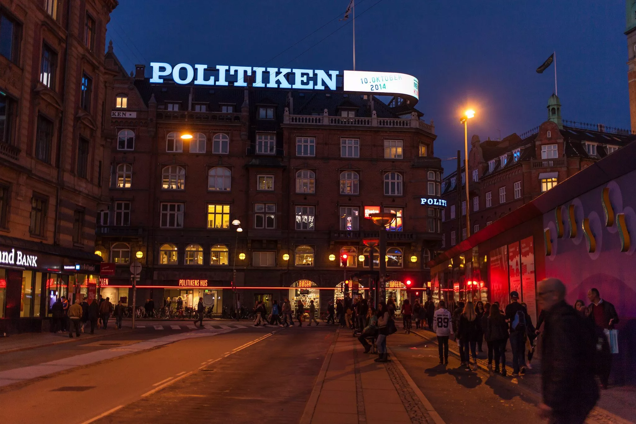 En bygade i skumringen med gående og siddende mennesker. Bygningen i baggrunden er oplyst og har et stort POLITIKEN-skilt på taget. Gadelygter og oplyste vinduer giver scenen en varm glød.