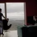 To mænd befinder sig i en moderne stue med store vinduer med udsigt over et landskab. Den ene mand står ved vinduet og holder en stol, mens den anden sidder i en grøn sofa og kigger ud. Scenen er blødt oplyst med naturligt lys.