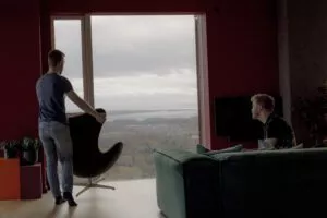 To mænd befinder sig i en moderne stue med store vinduer med udsigt over et landskab. Den ene mand står ved vinduet og holder en stol, mens den anden sidder i en grøn sofa og kigger ud. Scenen er blødt oplyst med naturligt lys.