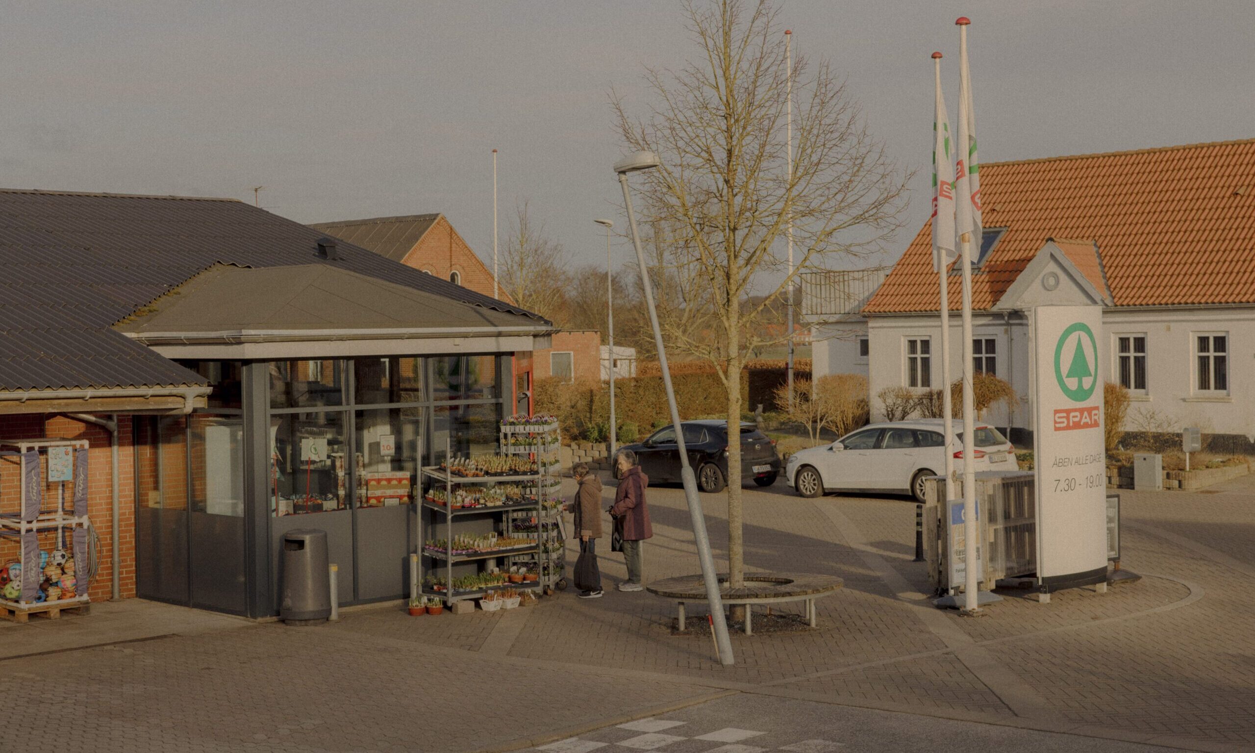 En lille købmandsbutik med et SPAR-skilt, udendørs planteudstilling, to mennesker, der taler sammen nær indgangen, parkerede biler og nærliggende bygninger på en solrig dag.