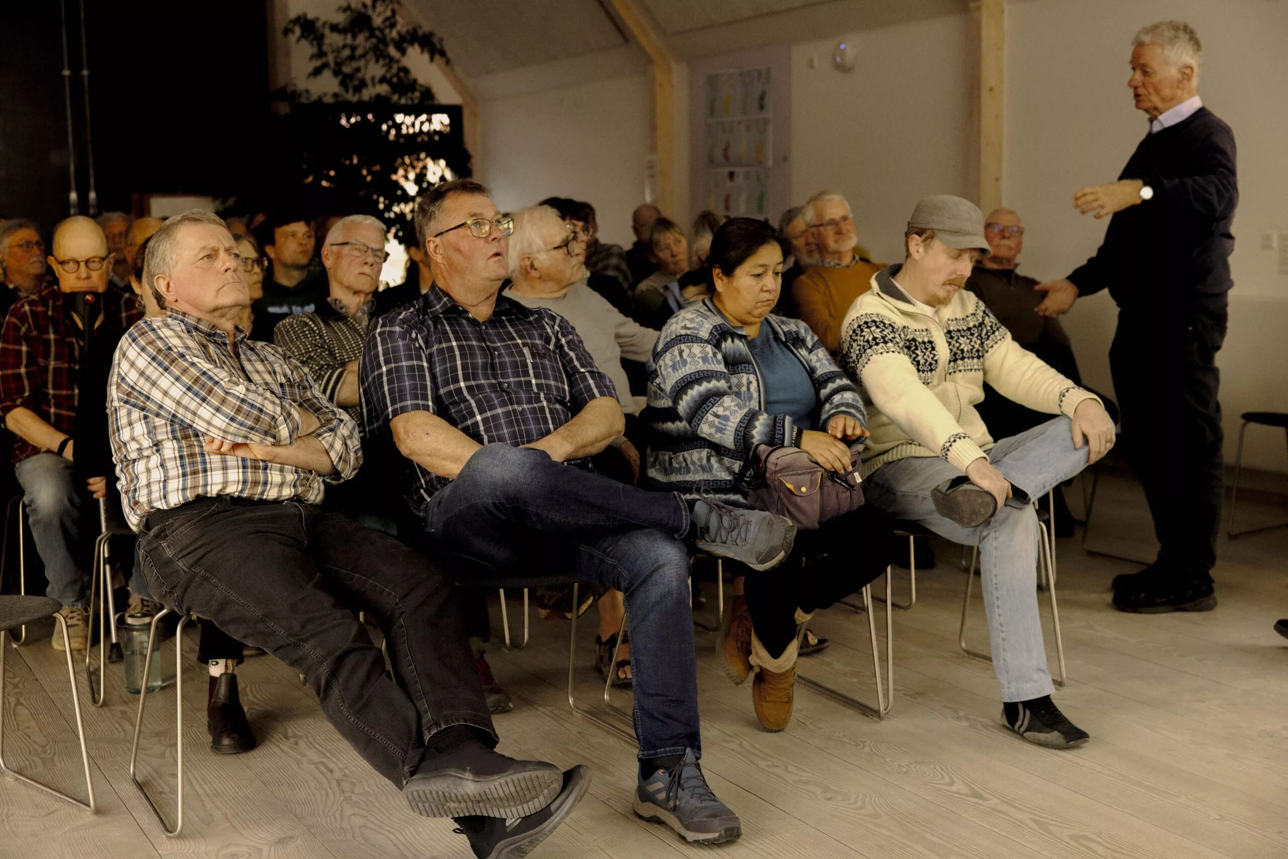 En gruppe voksne sidder opmærksomt på stole i et godt oplyst lokale og lytter til en mand, der står og taler foran dem. De fleste deltagere virker fokuserede, og nogle lægger armene eller benene over kors.