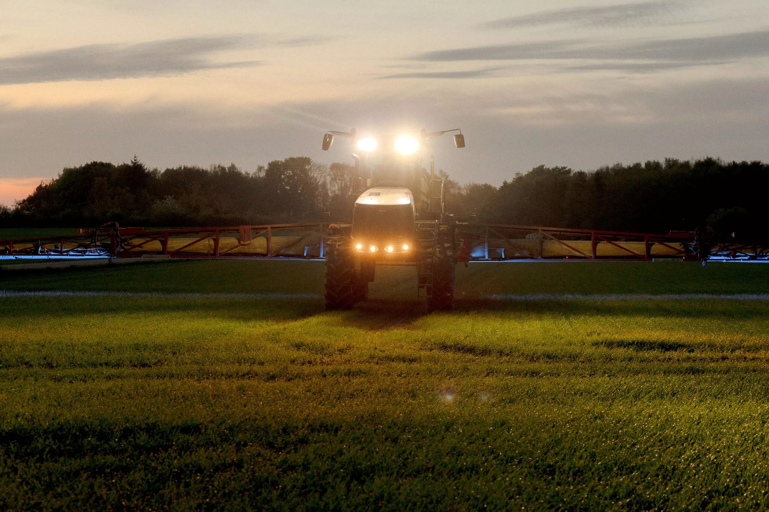 En traktor med forlygterne tændt sprøjter afgrøder på en mark i skumringen med træer og en overskyet himmel som baggrund.