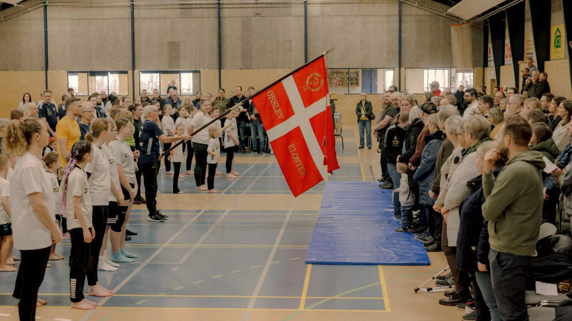 En gruppe mennesker står i en gymnastiksal, nogle i uniform, over for en person, der holder et rødt og hvidt flag. Tilskuere er samlet langs højre side og ser på begivenheden. Scenen ser ud til at være en formel eller sportslig ceremoni.