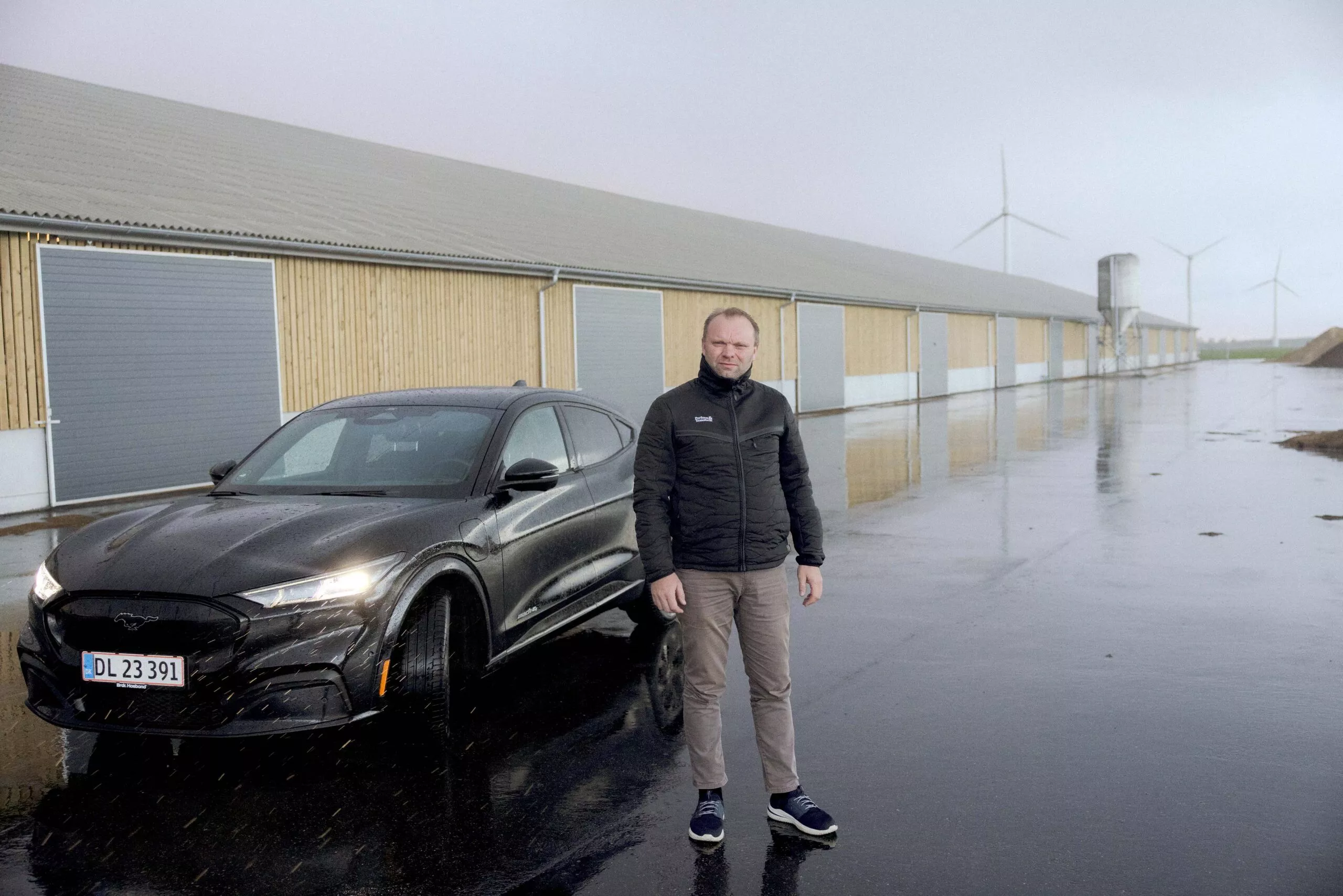 En mand står foran en sort bil på en våd, reflekterende overflade i nærheden af lange industribygninger. Vindmøller er synlige i baggrunden under en overskyet himmel.