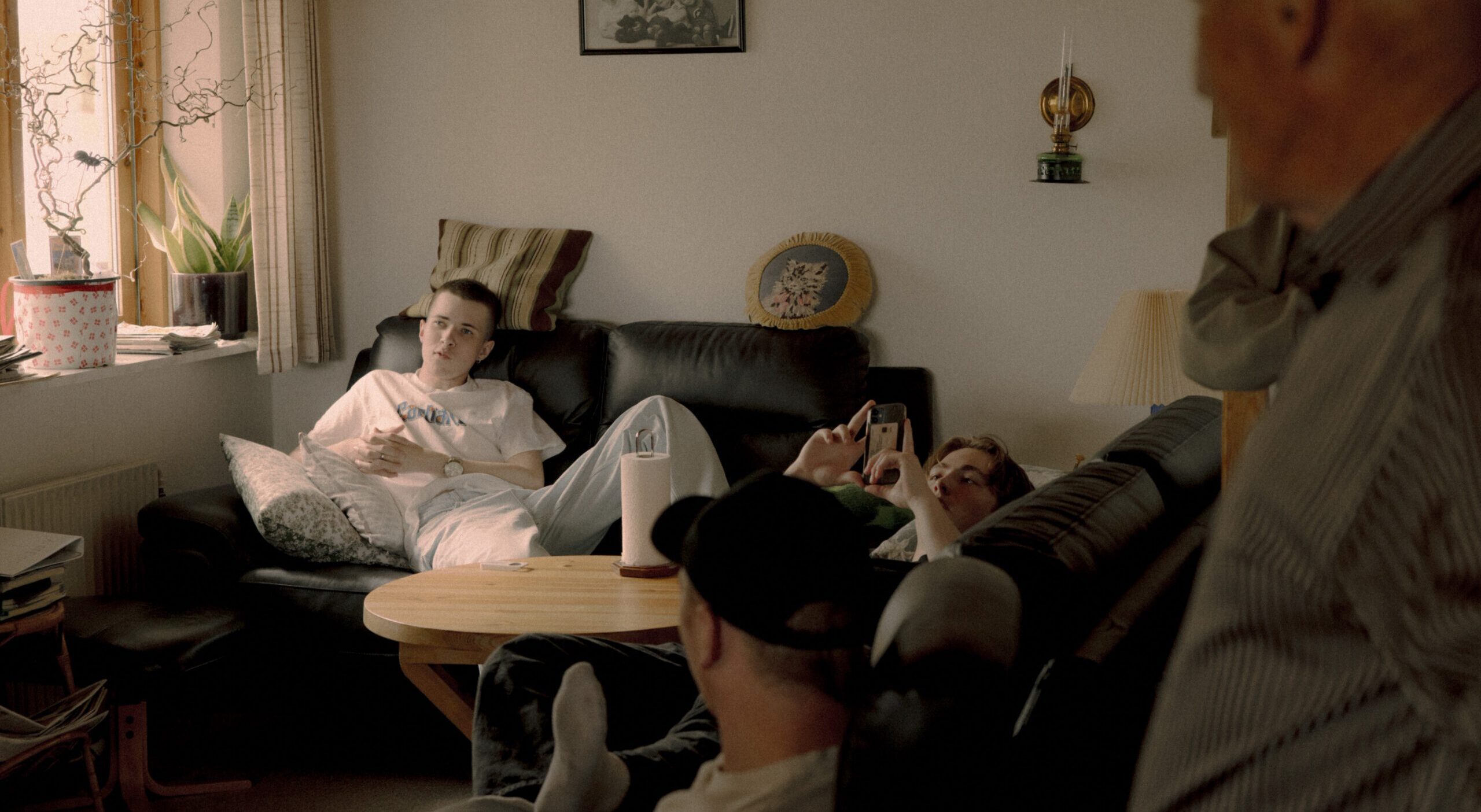 Tre unge mennesker slapper af på sofaer i en hyggelig stue, den ene bruger en telefon. En ældre mand står i forgrunden, delvist ude af fokus. Familiefotos og planter pynter i rummet. Varmt lys trænger ind gennem et vindue.