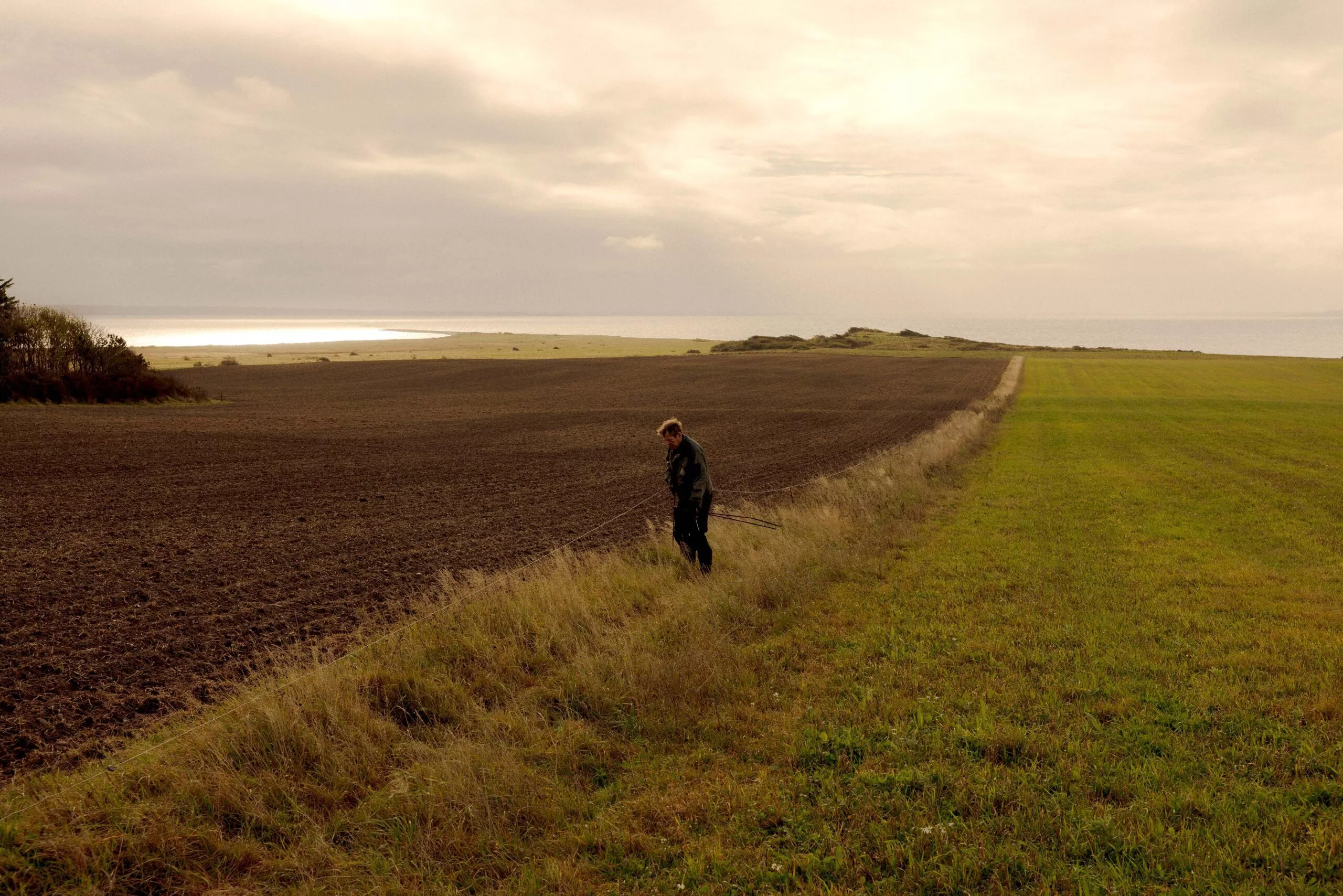 En person står nær kanten af en pløjet mark og et græsareal under en overskyet himmel og kigger ned med pinde i hånden; landskabet strækker sig ud mod fjernt vand og horisonten.