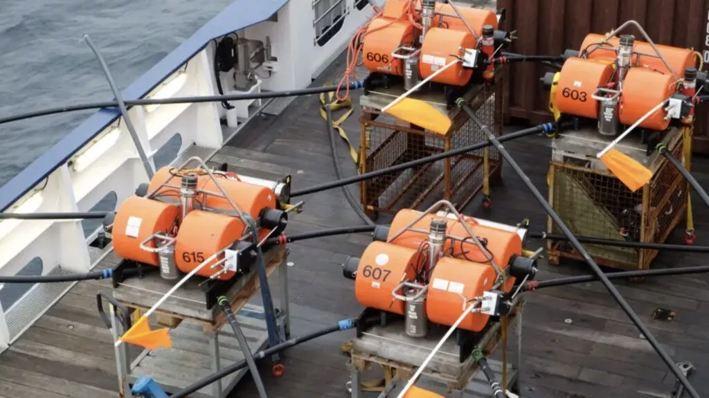 Flere orange videnskabelige instrumenter, mærket med numre, er fastgjort på metalstativer og træbure på et skibsdæk, omgivet af sorte kabler og orange flag, med havet synligt i udkanten.